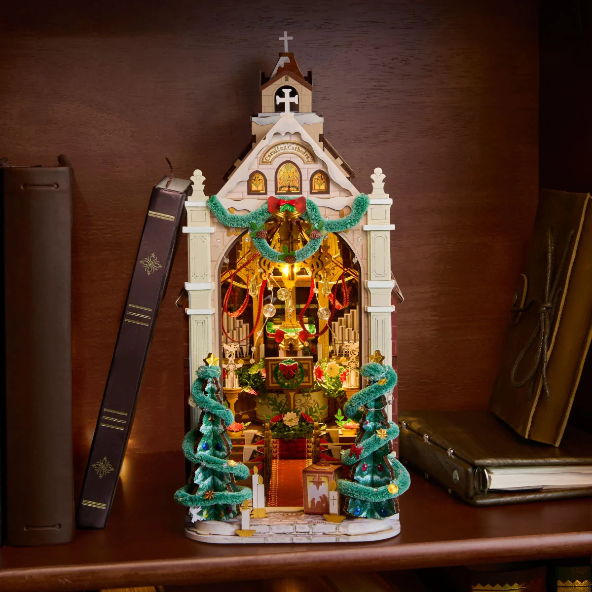 Rolife Caroling Cathedral illuminated Christmas book nook displayed on a wooden bookshelf between books as a festive miniature decoration